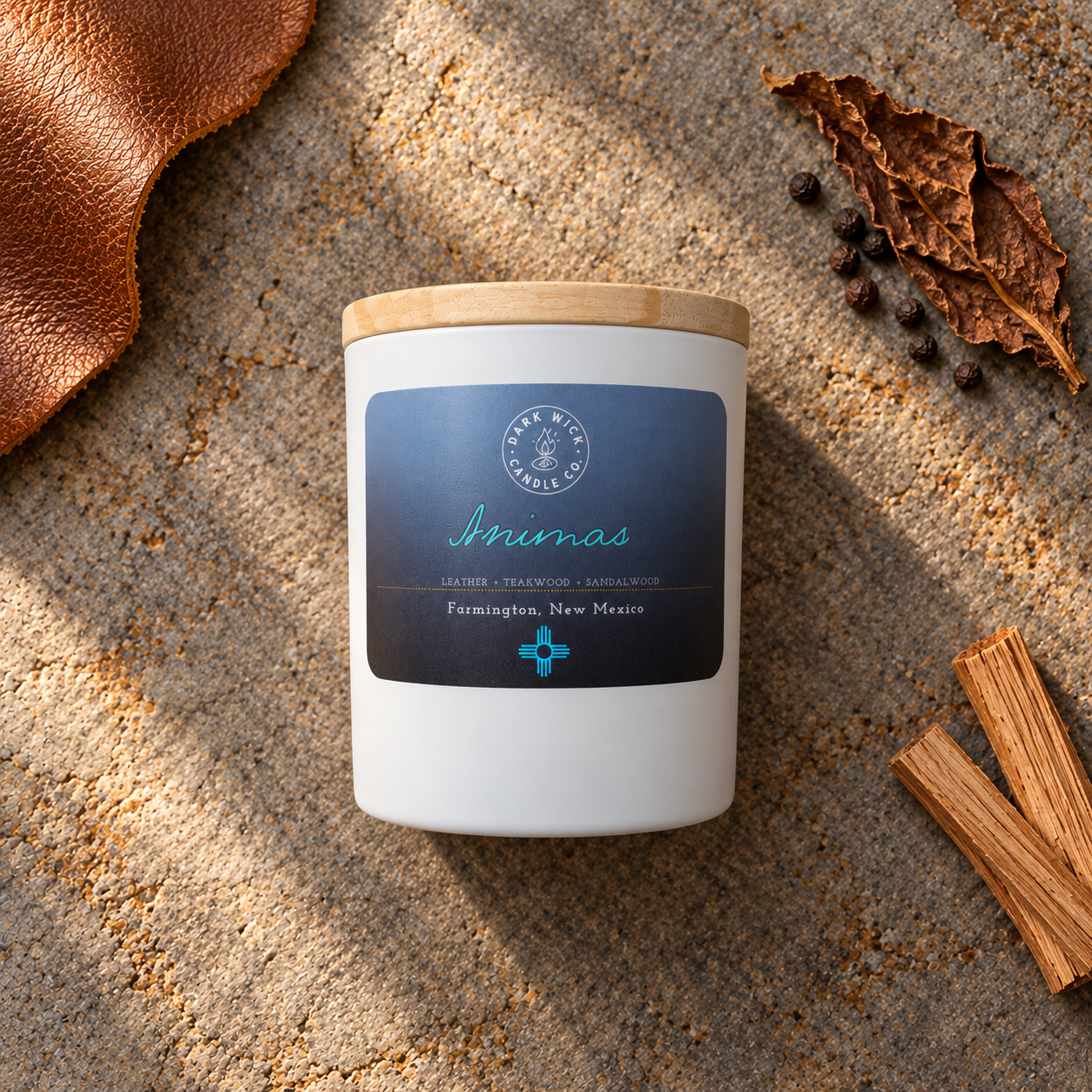 Flat lay of the small Dark Wick Animas candle in a white vessel with a wooden lid, surrounded by leather, a dried leaf, peppercorns and wood.