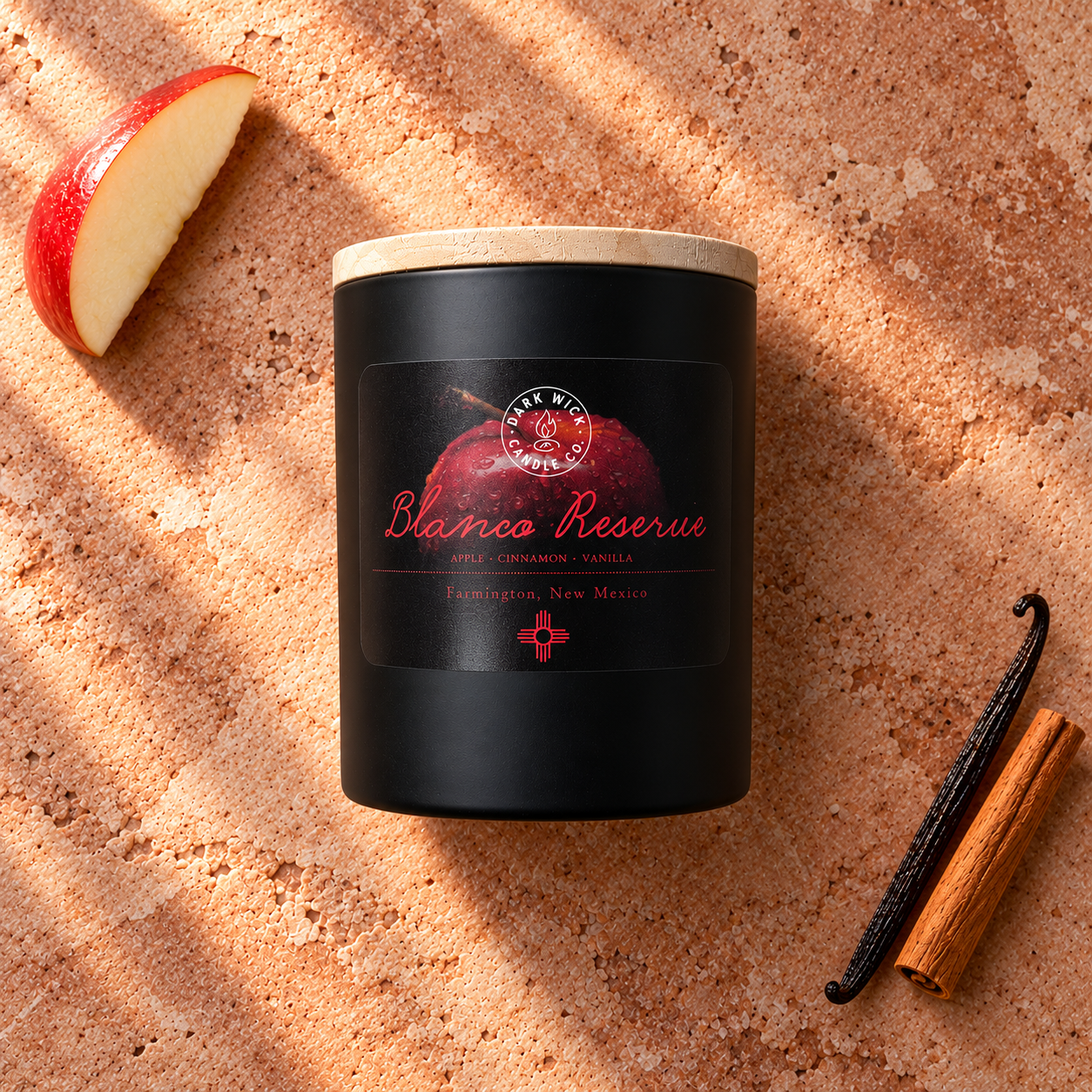 Flat lay of the small Blanco Reserve candle in a black vessel with a wooden lid, beside apple, cinnamon and vanilla on warm stone.