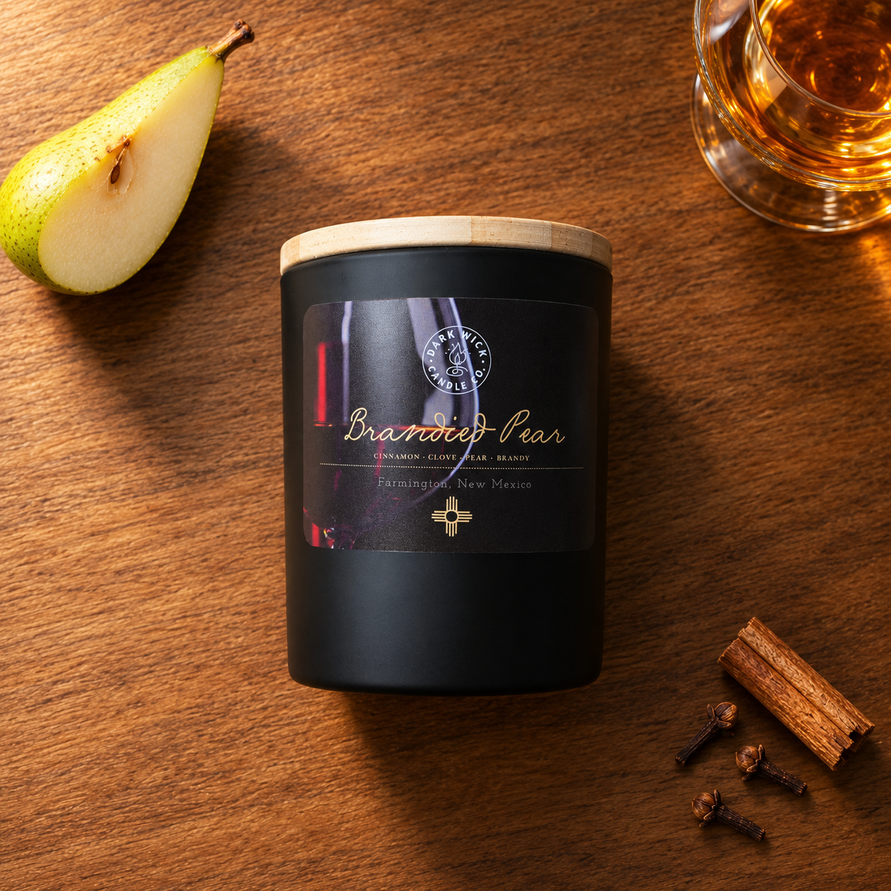 Flat lay of the small Brandied Pear candle in a black vessel with a wooden lid, beside pear, a glass of amber liquid, cinnamon and cloves on wood.