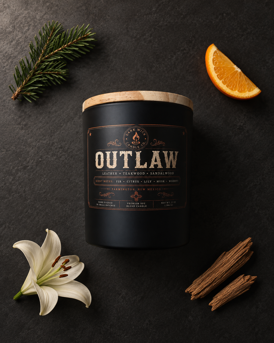Flat lay of the Dark Wick Outlaw candle in a black vessel with a wooden lid, beside citrus, fir, a white flower and wood on dark stone.