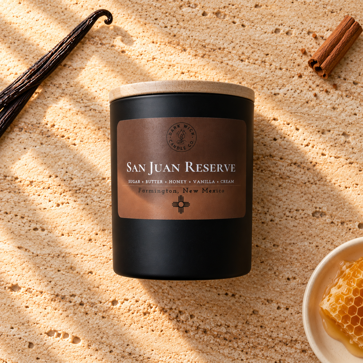 Flat lay of the small San Juan Reserve candle in a black vessel with a wooden lid, beside vanilla, cinnamon and honeycomb on warm stone.