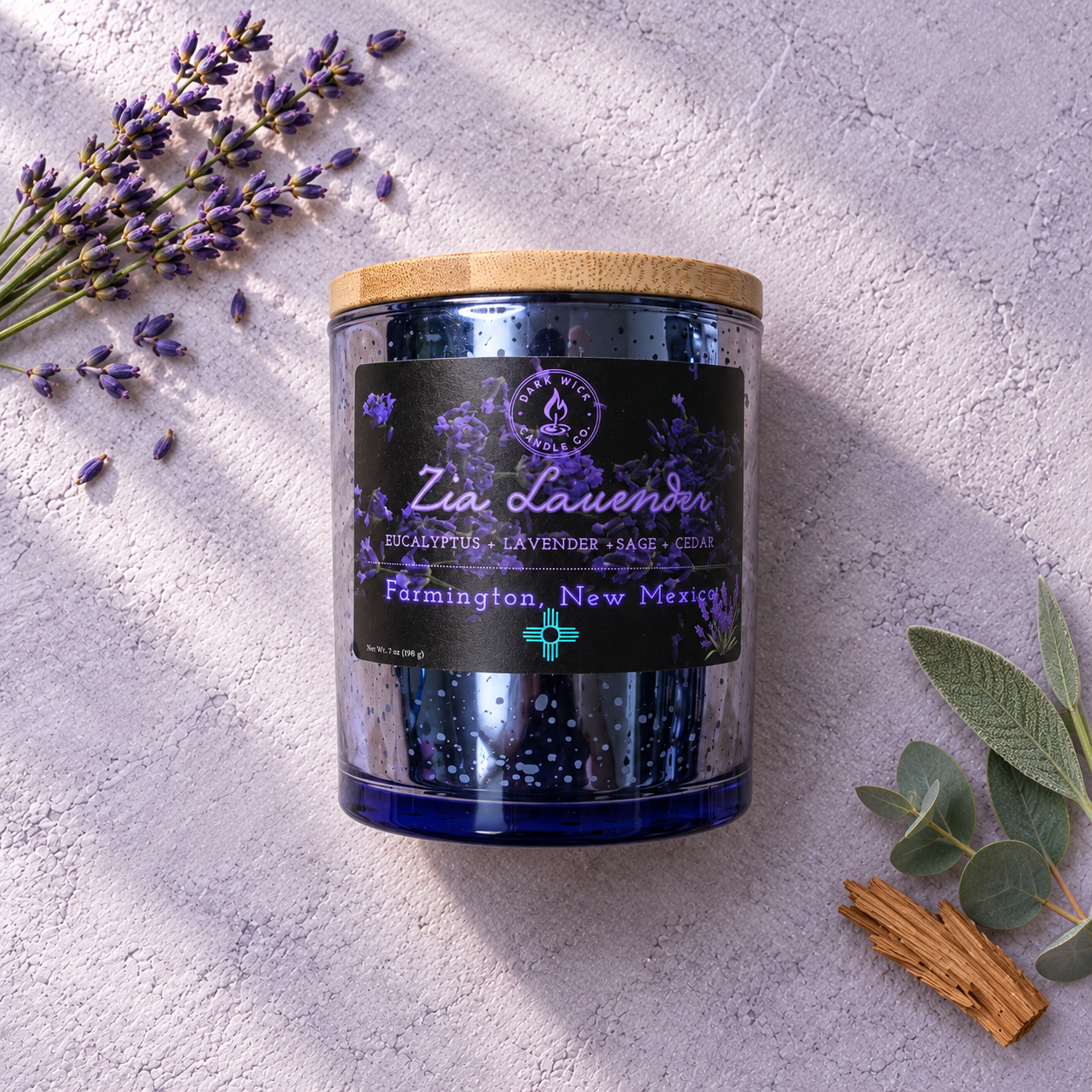 Flat lay of the small Zia Lavender candle in a blue glass vessel with a wooden lid, beside lavender, leaves and wood.